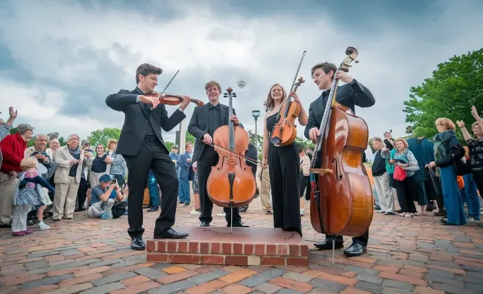 A dynamic photo of a student string quartet performing in an outdoor plaza, with a diverse crowd gathered around them, capturing the joy and engagement on both performers' and audience members' faces