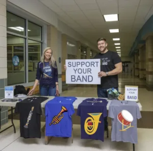 parents at a table selling band instrument themed tshirts to help raise money to buy instruments. The table is in the entryway of a highschool with the office and hallway in the background, Music Education